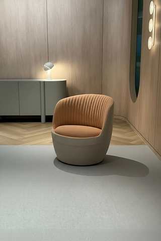 A modern interior scene featuring a light grey rug and a curved upholstered armchair in warm beige and terracotta tones, set against wood-paneled walls.