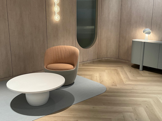 A modern lounge area featuring a ribbed peach-orange accent chair with a gray base, paired with a low white coffee table on a light gray rug. The room has light wood paneled walls, a matching wood herringbone floor, and a gray sideboard with a wall lamp.