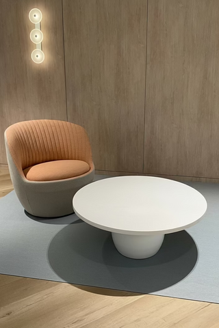 A modern living room setup featuring a round white coffee table and a curved armchair with a ribbed peach-orange fabric, set on a gray rug against a light wood-paneled wall. 